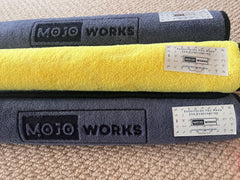 Three rolled branded Mojo Works surfcasting towels, two dark grey and one bright yellow, on a beige carpet