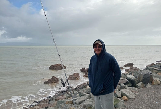 Man in blue hoodie and cap surfcasting on rocky shoreline with Mojo fishing rod near ocean under cloudy sky
