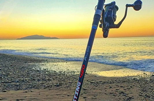 Mojo surfcasting rod with reel planted in pebbled beach sand at sunset, ocean and island view